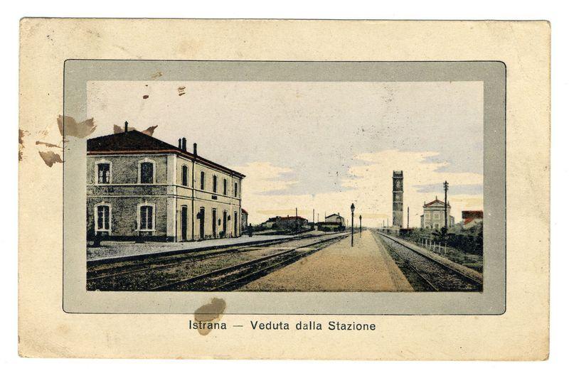 The image depicts a vintage postcard featuring an old railway station. The scene is framed by two sets of parallel railroad tracks extending towards the horizon, dividing the composition into three distinct sections: foreground, middle ground, and background.

In the foreground, there are several train cars on one track, suggesting activity within the rail yard or station area. These carriages have a vintage appearance with rounded windows typical of older trains.

The middle ground is dominated by an old two-story building, likely part of the railway infrastructure such as a maintenance shed or goods storehouse. The architecture includes multiple rectangular and arched windows, some boarded up which indicates disuse or abandonment in more recent times.

Behind this structure stands another taller building with classical features like columns at its entrance. This could be an important station facility or possibly even part of the main terminal building.

The background is mark