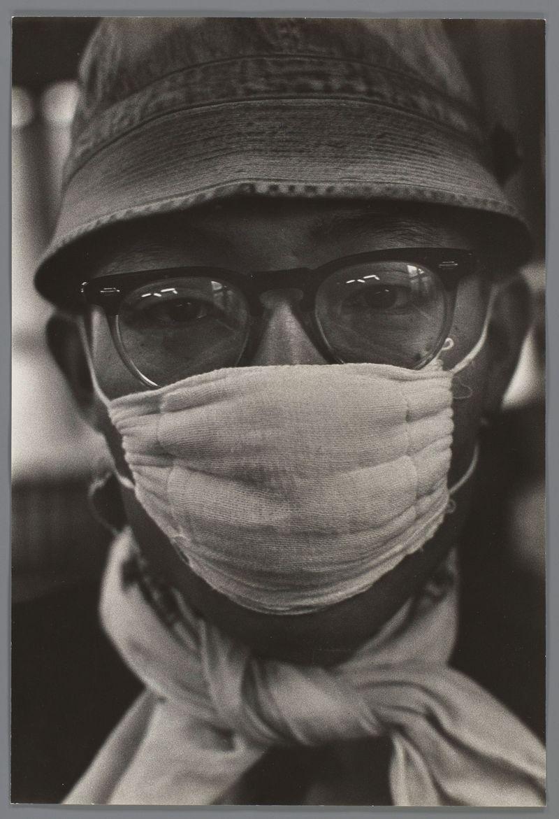 The image depicts a close-up of an individual wearing glasses and a face mask, creating the appearance that only their eyes are visible. The person is also dressed in what appears to be a hat or cap with some textural details on it. Below the chin area where the mouth should be, there's a cloth tied around the neck which contributes to a sense of privacy or anonymity. This style of photography was often used during times like war conflicts and pandemics as seen by contemporary works such "A Japanese Man With Face Mask" from 1959 by Ed van der Elsken where it reflects societal concerns about health, safety, identity concealment, and the blending in public spaces amidst a crisis. The grayscale tones of this photo highlight contrasts between light and shadow, focusing attention on facial expressions or elements that convey emotion or message without revealing personal details such as race, age, gender, etc., thus adhering to sensitive portrayal practices typical for many historical photog
