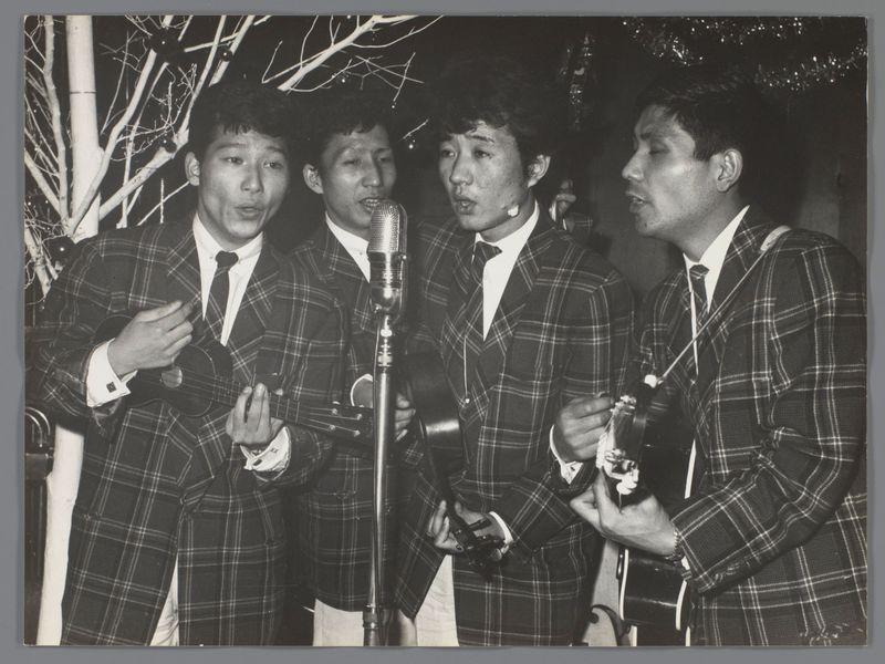 The image is a black and white photograph depicting four men on stage, likely performing as part of an amateur or semi-professional singing group. They are dressed in matching plaid suits with dark ties, suggesting they have coordinated their outfits for the performance.

Each man holds a musical instrument: three play ukuleles while one appears to be leading by speaking into a microphone, possibly introducing songs or engaging with the audience. The background is dimly lit and features bare tree branches, which could indicate an outdoor setting during winter or early spring nights. Above them hangs what seems like tinsel or garland decorations, adding a festive atmosphere.

The men's expressions vary from focused to expressive as they engage in their performance. This group appears united by camaraderie and the shared passion for music. The image captures a candid moment of cultural expression within Japan during the post-war era, reflecting on leisure activities among Japanese ci