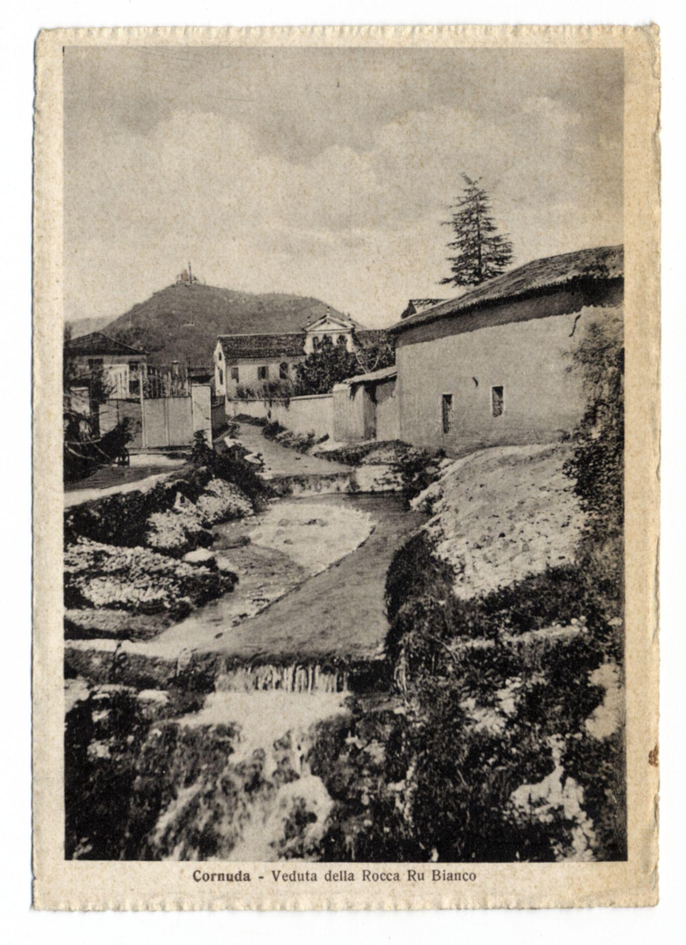 The image depicts a historical postcard featuring an old, rural scene. It shows a small stream meandering through rocky terrain and leading towards the foreground of the picture where it appears to flow into or out from behind vegetation. On either side of this waterway are structures resembling traditional European farmhouses with whitewashed walls, dark roofs, and some patches that suggest wear over time, indicative of age.

In the background, a hill is visible with what seems like ruins on top - possibly remnants of an old structure or fortification, which could explain the mention of "Rocca" in the caption. The architecture includes simple designs without ornate details, suggesting practical usage rather than display purposes.

The vegetation appears sparse and typical for arid regions; a tall tree stands prominently near one building. There is no visible human activity, lending to an atmosphere of stillness or abandonment. Above this pastoral scene are faint clouds in the sky,