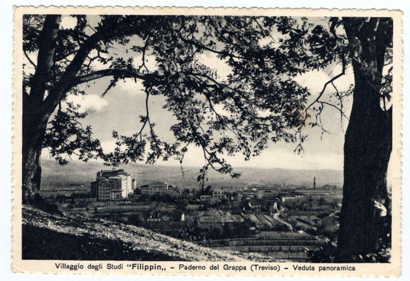 The image displays a vintage postcard depicting an aerial view of the Paderno del Grappa village in Treviso, Italy. It appears to be taken during daylight with ample cloud cover, casting soft shadows on the landscape below.

In the foreground, there is a prominent natural setting featuring two trees framing the left and right sides of the frame, their branches extending into the sky above. The foliage suggests it may have been captured in late spring or summer due to its dense appearance.

The middle ground reveals an urban area with multiple buildings spread out across what seems like hillsides. Notable among these are larger structures that could be residential complexes or possibly a religious institution given their architectural prominence and size relative to the surrounding houses.

In the background, there is an expansive view of flatlands leading towards another range of distant mountains under a pale sky indicative of either early morning or late afternoon light conditi