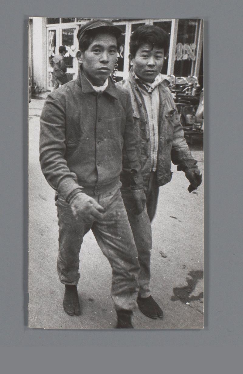 This black and white photograph captures two men walking on a street. They are dressed in work clothes, indicating they might be involved in manual labor or construction tasks. The man on the left wears a dark jacket with buttons, pants tucked into boots, and is wearing gloves while holding something small in his hand. His facial expression is neutral as he looks directly at the camera. The second man has short hair and appears slightly out of focus behind him; his attire includes a lighter-colored jacket that seems to be worn over another garment, possibly for warmth or protection from the elements.

The setting suggests an urban environment with shops in the background where people are visible through glass windows. There's no clear indication of their exact location within Japan due to language and cultural symbols being absent. The overall feel is candid, capturing a slice-of-life moment without posing or staged interaction between subjects and photographer.