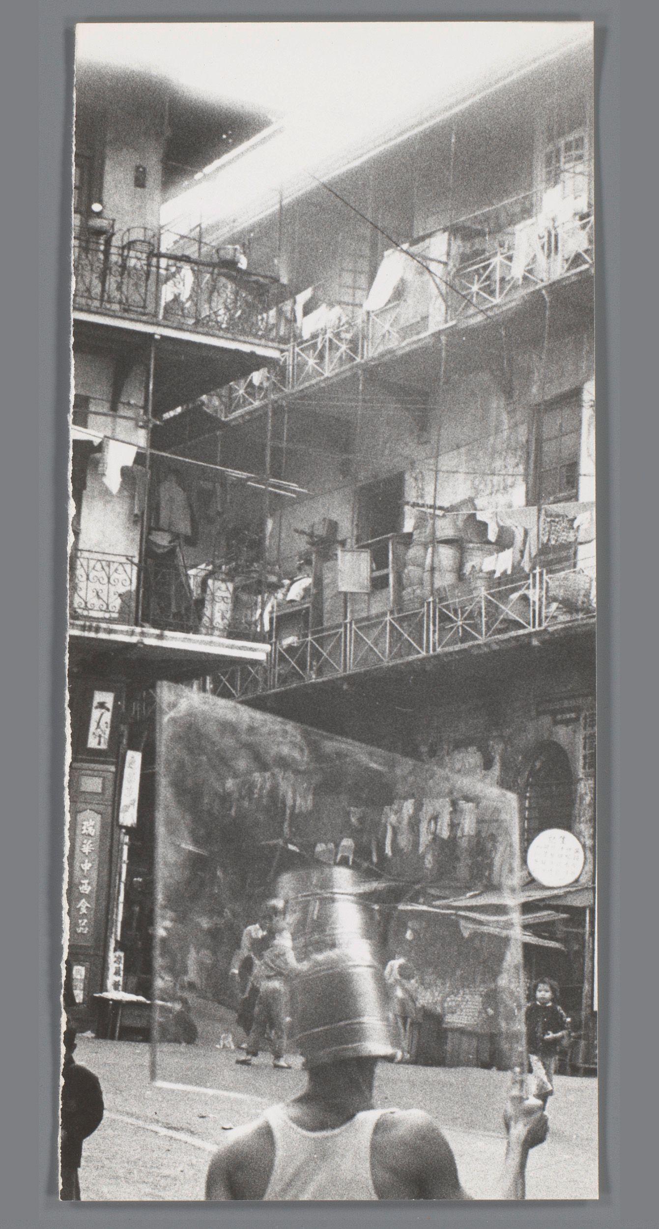 The image displays a black and white photograph, seemingly from the mid-20th century, capturing an urban street scene. In the foreground, there's a person wearing what appears to be a metal bucket on their head, suggesting they may have been engaged in manual labor or water collection. This figure is seen walking away from the viewer down the street.

The background reveals multiple floors of buildings tightly packed together, indicative of densely populated urban housing. Various individuals are visible on these balconies and within doorways, going about daily activities such as hanging clothes to dry. The architecture suggests a somewhat disheveled environment with exposed pipes and laundry lines strung between structures.

Visible signage indicates the presence of businesses or restaurants in this residential area. One sign reads "RYUJI'S CHINESE FOOD," suggesting an establishment serving Chinese cuisine, which aligns with Japan's historical connections to China as well as Japan