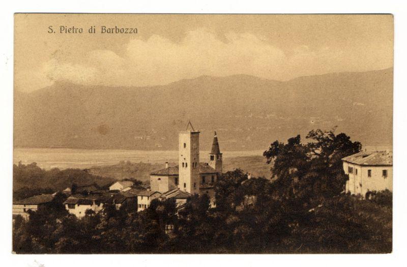 The image depicts a vintage postcard showing an old village named S. Pietro di Barbozza, possibly from the early 20th century given its sepia-toned appearance and style of photography. In the foreground are dense clusters of trees with some buildings peeking through them. The middle ground is dominated by two prominent church towers rising above a cluster of houses; one tower has rectangular windows while the other features circular ones, suggesting different architectural styles or periods for each building. Further back, more residential structures and a body of water can be observed under a cloudy sky. In the far background, mountains loom over the landscape. The postcard's title is prominently displayed at the top in bold uppercase letters against the faded image backdrop.