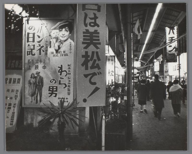 This black and white photograph captures a bustling street scene in Japan, likely from the late 1950s to early 1960s. The focus is on several large vertical banners with Japanese text and some English writing, which suggest they are advertisements for various products or events.

One prominent banner features an image of two people, presumably actors Anne Frank and someone else, promoting a film titled "The Diary Of." Other visible texts include phrases like "松" (which means pine) and other characters that likely denote names or titles related to the promotions. The style of clothing worn by individuals in the photo indicates it is not contemporary.

Pedestrians are seen walking on the street below these banners, with their movements blurred slightly due to a long exposure setting used when taking the photograph. This technique gives them an impression of motion, suggesting the vibrancy and pace of life at that time. The lighting appears artificial, typical for urban nighttime sett