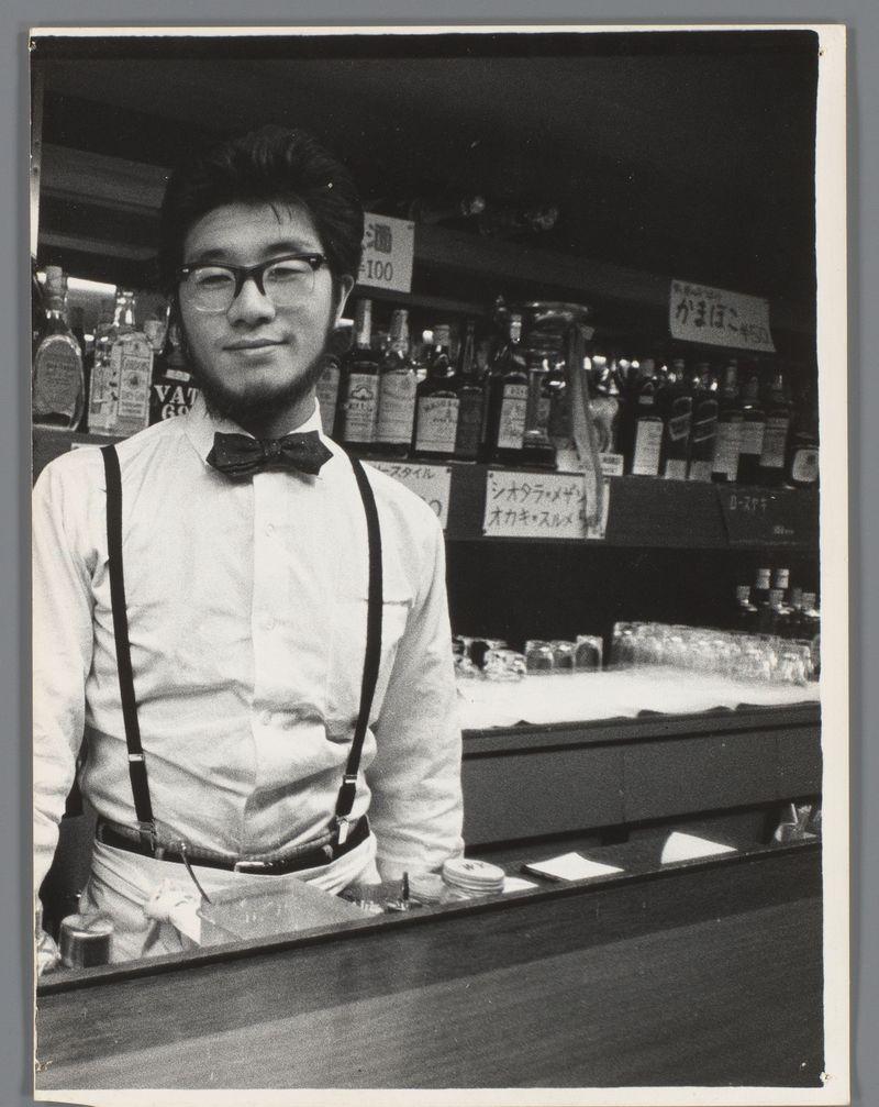 The image depicts a black and white photograph of an individual who appears to be bar staff, standing behind a counter in what looks like a bar or restaurant setting. The person is wearing glasses, a bow tie, suspenders over the shoulders, and has a neat hairstyle typical for that era's fashion sense. In the background on shelves above the counter are various bottles labeled with prices and Japanese characters, indicating an Asian locale likely from Japan given historical context.

The individual appears to be in their mid-twenties or early thirties, suggesting they could have been quite young at the time of this photograph which is dated between 1959-60. The attire and setting suggest a formal yet approachable working environment typical for that period's service industry professionals.
