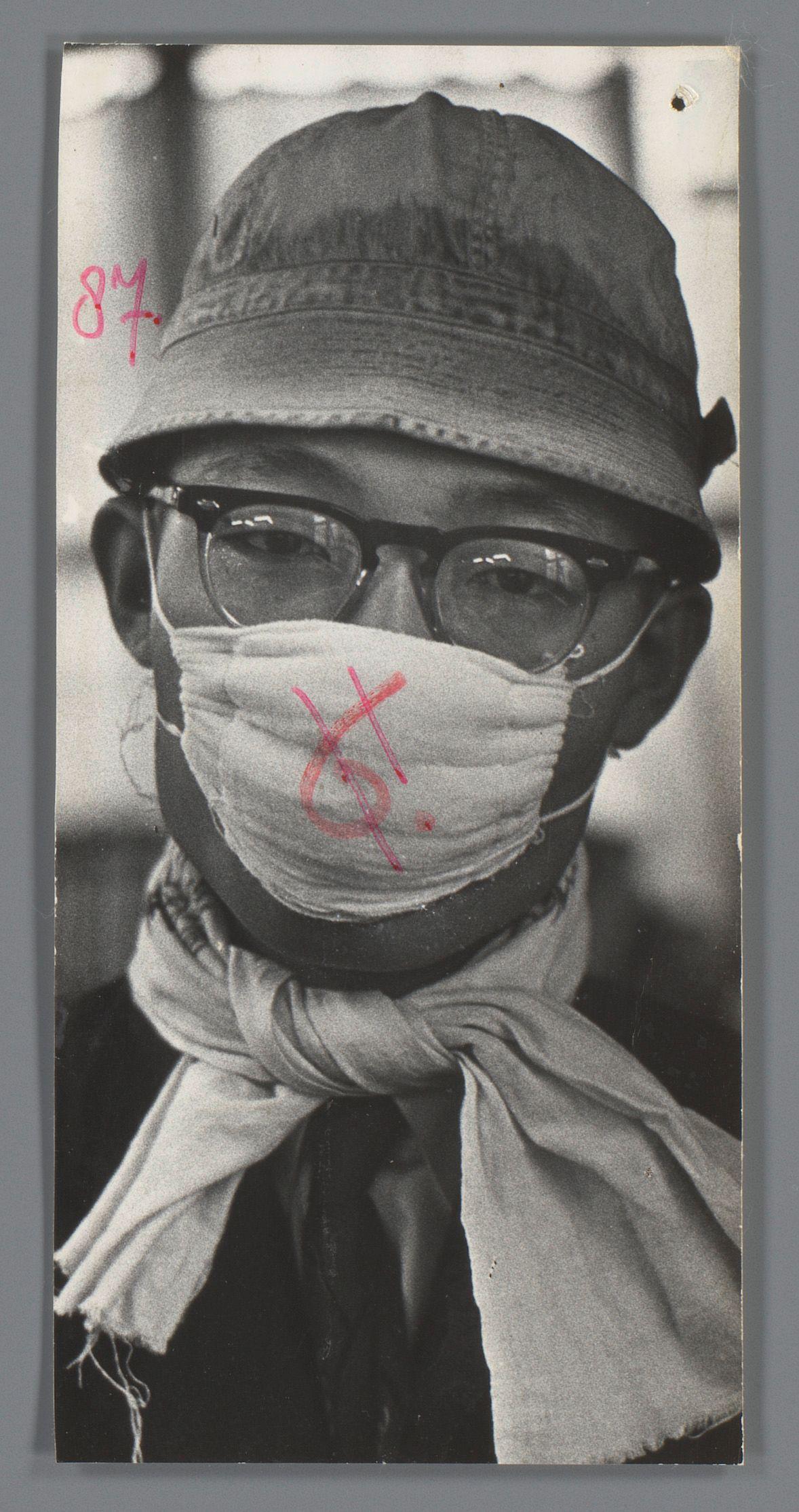 The image depicts a person wearing glasses, with their face partially obscured by a medical mask and scarf. The individual is also seen wearing a cap or hat that appears to be made of fabric, possibly cotton. The photograph has a monochromatic appearance, predominantly in shades of gray, suggesting it may have been taken during the 20th century, potentially related to historical contexts such as wartime measures or public health concerns like pandemics.

The mask is notable for having red marks on it, which might indicate some kind of notation or mark made after the photograph was captured. The person's expression and posture are not fully visible due to the face being covered by the mask and scarf, adding a layer of mystery to their identity and intentions at the time when the photo seems to have been taken.

The image carries historical significance as it could reflect the conditions during specific periods in history where such preventive measures were common. The presence of wh