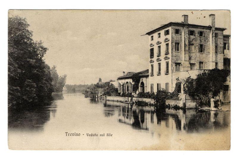 The image depicts an old, sepia-toned postcard showing a picturesque scene of a canal or river running through what appears to be the historic center of Treviso. On one side of the waterway stands a large building with multiple windows and architectural details characteristic of classical European design, possibly from the 19th century given its style. The reflection on the calm surface of the water adds symmetry to the composition.

Trees line the banks of the canal or river, suggesting an urban environment that integrates nature within city limits. There is a faint figure in white near the right edge of the postcard by the water's edge, possibly indicating human presence and activity from this time period. The overall atmosphere evokes nostalgia for past times, with a sense of quietude conveyed through the lack of visible movement or crowds.

At the bottom left corner, text reads "Treviso - Veduta sul Sile," which translates to "Treviso - View on the Sile River." This indicates t