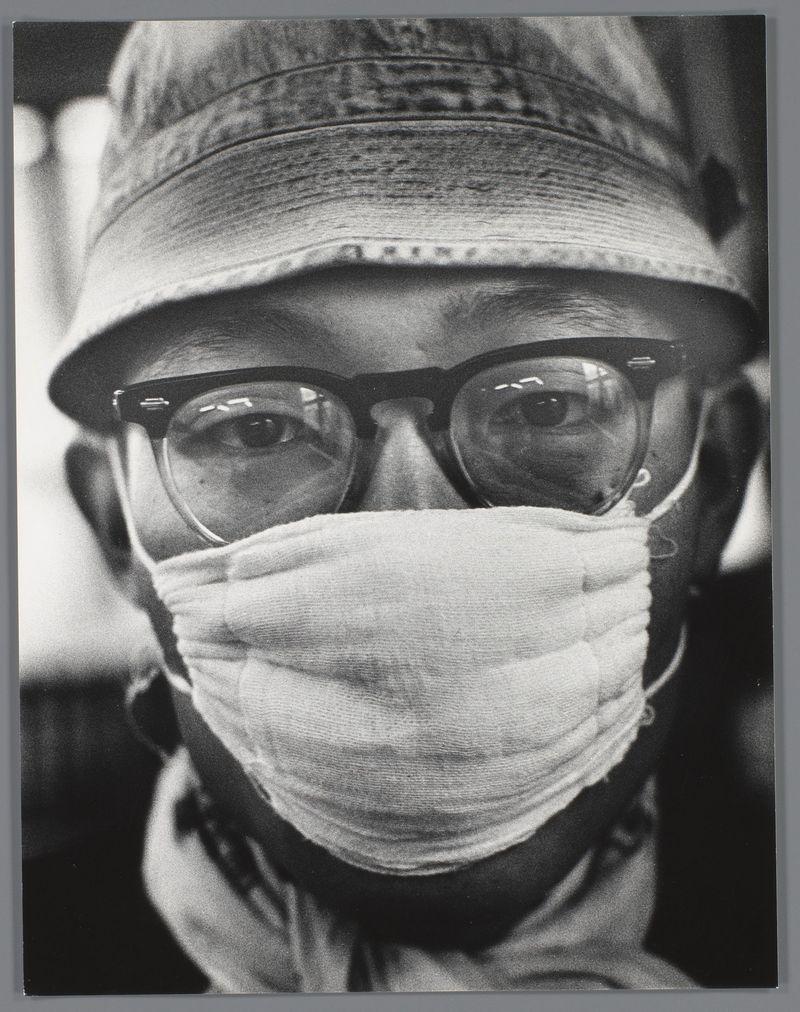 The image is a black and white photograph of an individual wearing glasses, with the lower half of their face obscured by a light-colored surgical mask. The person appears to be older, as suggested by the visible lines on their forehead and around the eyes within the eyeglass frames. They are also wearing what seems to be a cap or head covering that is wrinkled in texture, which could indicate movement during photography.

The background of the photo is blurred, drawing focus towards the individual's face. The lighting creates strong contrasts between light and shadow on their features, particularly around the eyes framed by glasses. There is no discernible context or setting visible to provide additional information about where this image was taken or what it represents beyond its subject.