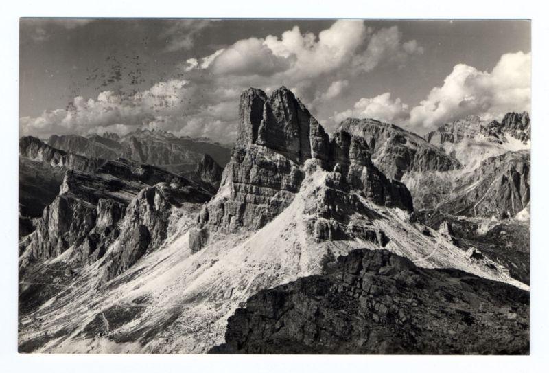 The image shows a dramatic and rugged mountain landscape captured in black and white. In the foreground, there is an imposing rocky peak with sharp, jagged edges that dominate the central portion of the frame. This main summit has two distinct peaks rising sharply from its base, creating a natural arch-like formation known as "Ferro Candilera" which translates to "Iron Candlestick." Surrounding this prominent feature are other rock formations and cliffs showing various textures and shapes.

The background is filled with an expansive view of the mountain range extending into the distance. These distant peaks appear softer in contrast, suggesting a sense of depth and scale. The sky above features large clouds that add dynamic movement to the scene, creating areas of light and shadow across the landscape.

Overall, the image conveys a powerful portrayal of nature's majesty through its monochromatic tones and composition focused on verticality and sharp contrasts between different elem