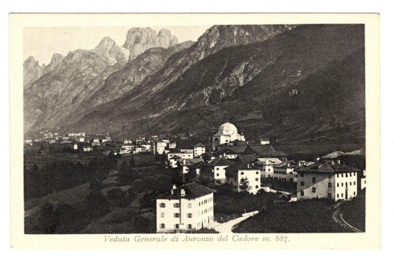 The image is a black and white photograph, seemingly from the late 19th century or early 20th century given its style. It depicts a general view of Auronzo del Cadore as seen in m.887. The landscape prominently features towering mountain ranges with sharp peaks that dominate the background. In contrast to this rugged natural scenery are several buildings nestled within valleys and on slopes, indicating human habitation or settlement.

Notably highlighted among these structures is a large dome building which stands out due to its size and distinctive architecture, suggesting it may be of religious significance such as a church or cathedral. The town's layout shows relatively spaced-out development with pathways leading through the residential areas towards what seems like a central point where one could gather.

The photograph has text at the bottom that reads "Veduta Generale di Auronzo del Cadore m, 887," indicating it is likely part of a series or catalog showing different views 