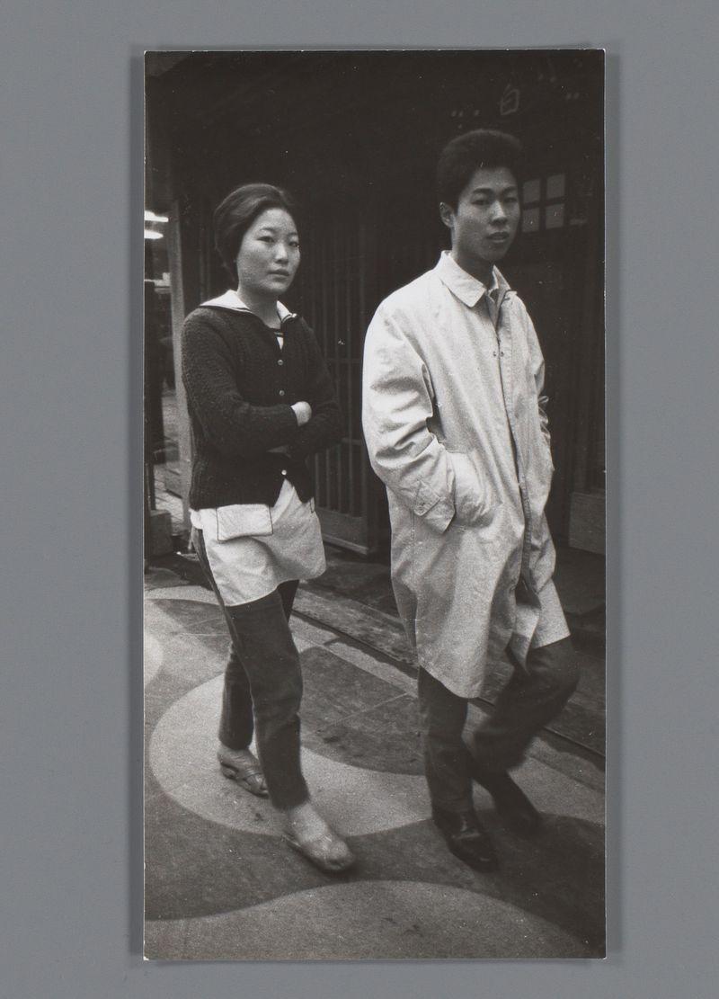 The image depicts a young Asian man and woman standing on what appears to be a street or sidewalk. The photo is in black and white, suggesting it could be from an earlier time period. Both individuals are dressed casually; the man wears a light-colored long-sleeve shirt and dark pants with boots, while the woman has her arms crossed, wearing a dark sweater over a lighter top paired with darker bottoms and shoes that look like sandals or flip-flops.

The background is somewhat blurred but shows an urban setting with architectural elements indicative of buildings. The street appears to be made up of large blocks, possibly cobblestone or paving stones commonly found in older city centers.

There's no specific context provided for the photograph; it captures a candid moment between two individuals on a public space without any clear indication of their relationship or purpose for being there.