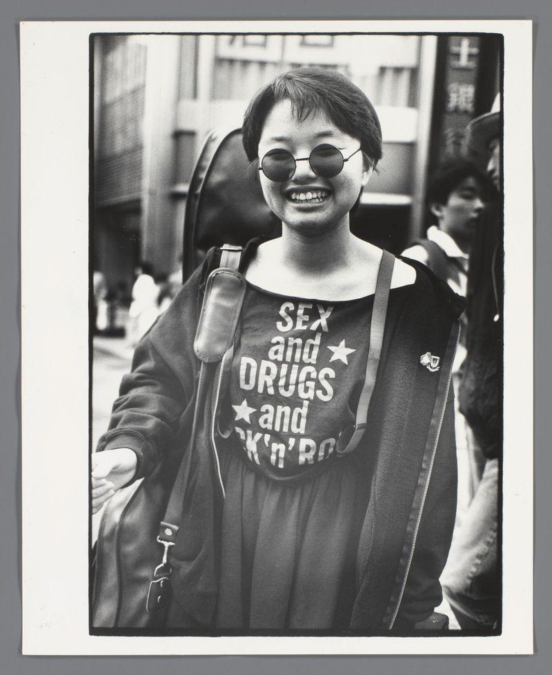 The image is a black and white photograph featuring an Asian woman with glasses smiling at the camera. She wears a casual outfit consisting of dark clothing, including what appears to be a dress or jacket over her shoulders, along with sunglasses on her face. The most striking aspect of this photo is her T-shirt adorned with bold lettering that reads "SEX AND DRUGS and ROCK 'N' ROLL" in white text against a black background, punctuated by stars around the word "DRUGS." This attire suggests she may be attending or participating in an event related to music culture. The woman carries a shoulder bag on her left side as she stands among other individuals who are partially visible in the blurred urban backdrop. Her joyful expression and distinctive outfit make this image memorable, captured by photographer Ed van der Elsken during his travels in Japan around 1988.