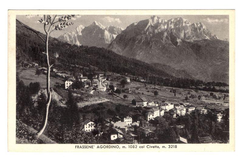 The image is a vintage black and white photograph depicting the village of Frassanè' Agordino, with an elevation marked as 1.082 meters at Col Civetta, which measures approximately 3.218 meters. The scene showcases a picturesque landscape where a small settlement nestles against rolling hills that rise into majestic mountain peaks in the background. Prominent among these mountains is one known colloquially as 'Civetta,' suggesting its significant stature and possibly an association with local folklore or geography.

The village consists of several white-walled buildings, indicative of traditional alpine architecture, spread out across a valley floor dotted with cultivated fields and trees. A few solitary structures stand at the foreground on sloping terrain leading towards the central cluster of dwellings, while more homes sprawl upward along paths that twist through the landscape.

A prominent tree stands alone in the lower left corner, perhaps symbolizing resilience or serving as a local landmark within this rural setting. The photograph's grainy texture and monochromatic palette suggest it was taken during an earlier era when such imagery captured daily life before modern digital photography became prevalent.