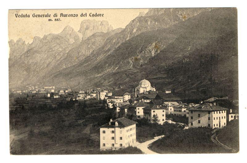 The photograph shows a general view of the town Auronzo del Cadore, as indicated by both Italian and German texts at the top. The text suggests that this image is part of a collection or archive with an identification number m. 887. In the center background lies a mountain range featuring rugged peaks, suggesting an alpine location typical for Italy's Dolomites region where Auronzo del Cadore is situated.

In the foreground and middle ground are various buildings characteristic of European architecture from past centuries; they include simple to medium-sized structures with pitched roofs, windows, and doors. The architectural styles vary slightly but collectively indicate a residential area possibly from an earlier time period given their design and condition.

The town seems compactly built around a central axis which could be considered the main street or path leading towards what appears as a domed structure in the center-right of the image; this building is distinct, larger tha