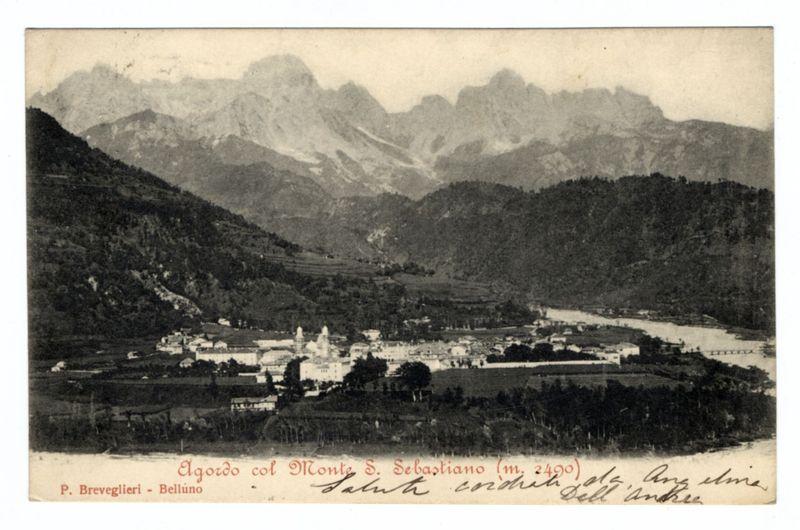 The image is a black and white photograph depicting a historical town nestled among the mountains. In the foreground, there are buildings closely packed together with visible rooftops but no discernible architectural details due to distance or resolution limitations. The midground features what appears to be an open space, possibly a field or public area adjacent to structures that could serve as market stalls or communal spaces.

The background is dominated by towering mountain peaks shrouded in mist and partially covered with snow, indicating the region's high altitude. A river meanders through the landscape near the town, adding a natural element to the scene.

Atop these mountains looms Monte S. Sebastiano at an elevation of approximately 2490 meters as noted by handwritten annotations on the photograph. The text indicates that this image is from Belluno and was taken with Monti S. Sebastian (m. 2490), suggesting a historical context where photographs were likely used for trave