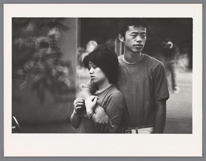 The image is a black and white photograph featuring two individuals standing closely together. The person on the left appears to be wearing a short-sleeved shirt with what seems like fur trim at the ends of sleeves, paired with dark pants or possibly shorts. This individual has shoulder-length hair and their arms are slightly raised in front of them. They appear focused or contemplative.

The second person is standing behind partially obscuring the first individual's right side while resting an arm over their shoulders. The visible portion shows this person wearing a short-sleeved shirt with what looks like belt loops at the waist, suggesting similar attire to the other figure but in darker colors that blend into the shadows of the background.

The backdrop is out-of-focus, indicating motion or low light conditions, and features indistinct shapes which could suggest an urban environment. The figures' expressions are not clearly visible due to their positioning within the frame; how