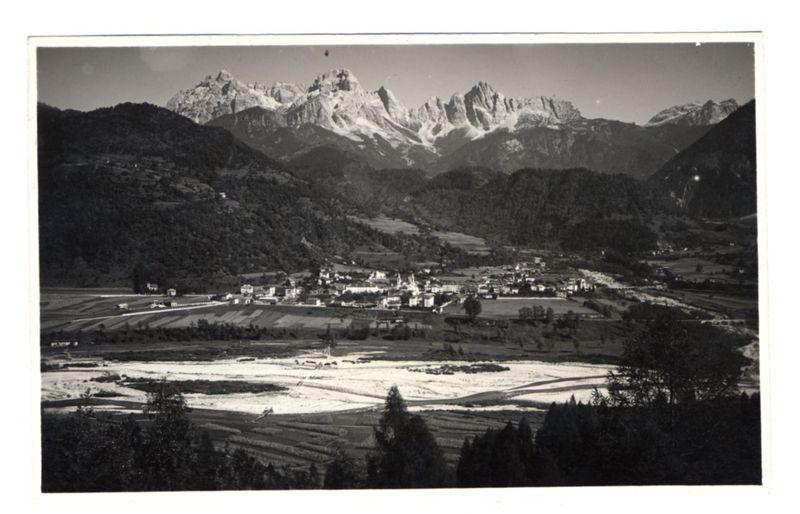 The image is a black and white photograph depicting a picturesque landscape dominated by snow-capped mountains. In the foreground, there's an expansive view of agricultural fields with patches that suggest different types or stages of cultivation, possibly rice paddies given their shape and patternation. The middle ground features what appears to be small settlements with clusters of buildings that could include homes, farms, or rural infrastructure. These structures are relatively close together but have a scattered arrangement amidst the greenery.

In the background, majestic mountains rise against a clear sky, indicating this landscape is likely at high altitude and possibly within a valley given how they flank either side of what looks like a river flowing from left to right across the image. The mountain peaks show variations in shape with some jagged while others are more rounded, suggesting geological diversity.

Notably, there's an object resembling a flag or marker on top 