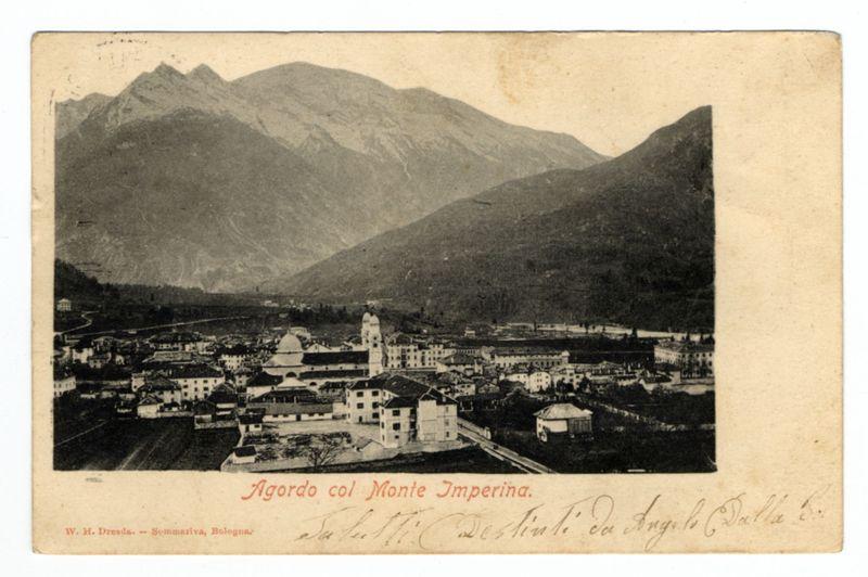 The image is a sepia-toned, aged postcard depicting the town of Agordo with Monte Imperina in the background. The mountains loom prominently over the settlement below. Architectural features include multiple buildings and structures that suggest European design, such as pitched roofs, chimneys, and what appears to be religious or government edifices due to their size and style. There is visible handwriting on the bottom right corner which could indicate a personal note or address related to this postcard's use.

The text "Agordo col Monte Imperina" in red script suggests that Monte Imperina is an important geographical feature of Agordo, possibly indicating its significance as part of a natural landscape view captured by the photographer. The presence of human signatures and inscriptions adds a personal touch, hinting at either ownership or gifting history for this postcard.

The overall composition conveys a sense of historical travel photography from perhaps late 19th to early 20