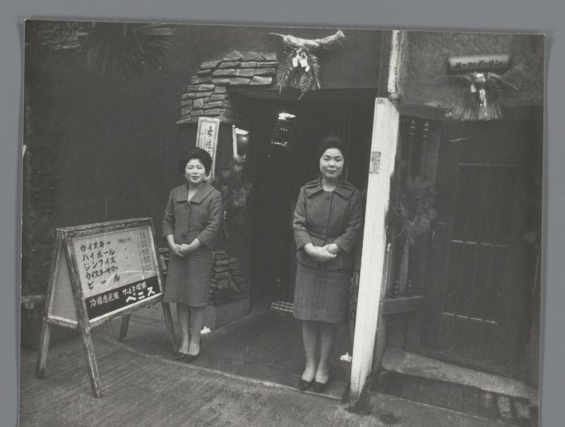 The image depicts two women standing in front of a café entrance. The setting suggests an older, perhaps mid-20th-century Japanese urban environment based on the architectural style and attire.

On the left stands a woman wearing glasses, a dark coat with a lighter patterned scarf or collar, and dark skirt. She has her hands clasped together at waist level while looking directly into the camera with a composed expression.

To her right is another woman in similar attire—a darker-colored striped coat over a matching dress—and she too gazes forward but with a slightly softer demeanor compared to the first.

They are positioned beside a door, which appears narrow and traditional. Above it hangs what seems like a straw hat or decorative piece tied together at the top, possibly part of Japanese cultural decor.

To their left is an A-frame signboard written in Asian characters—likely Japanese given context—and numerals indicative of prices, suggesting this café may offer food or drin