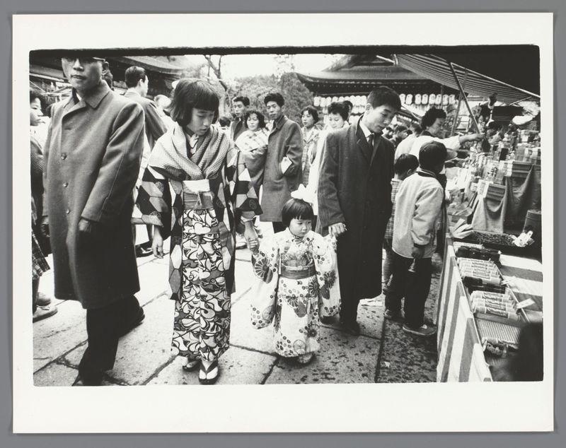 A black and white photograph depicting a busy market scene with numerous individuals browsing various stalls. The subjects are dressed in traditional Japanese attire, suggesting the photo was taken during a festival or special occasion. A child is wearing a decorative kimono while holding hands with an adult, possibly their parent, indicating family involvement.
