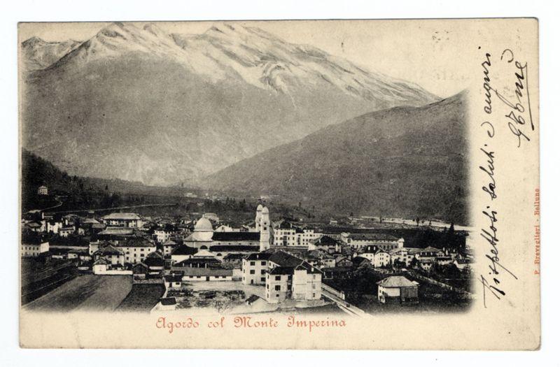 The photograph depicts a historical black and white image, possibly from the late 19th or early 20th century. It shows an elevated view of Agordo with Monte Imperina in the background. The town has traditional European architecture, including buildings with pitched roofs and some notable domed structures which suggest religious significance. In the foreground are open fields leading up to the urban area, while beyond that lies a vast expanse of mountains covered partially by snow or mist. There is handwritten text on the right side in what appears to be Italian script, alongside signatures at various places suggesting personal annotations possibly made after the original print's creation. The image carries an aged look with some discoloration and marks indicative of its age and perhaps handling over time.
There are no explicit indications of gender or race as it is a historical architectural photograph without human subjects present in focus.