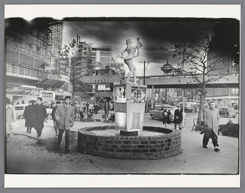 The image depicts a black and white street scene, seemingly from the mid-20th century. At its center is a prominent statue on top of an outdoor podium or pedestal with Japanese text indicating it may be in Japan, given to commemorate something significant.

Around this central feature are various elements that convey daily life: pedestrians walking by; individuals sitting around what appears to be maintenance work being done at the base of the sculpture. The background reveals urban architecture under construction and completed structures adorned with signage written in Japanese characters suggesting commercial establishments or businesses. Street furniture like lampposts, streetlights, and utility poles are visible.

The sky is overcast, casting a muted lighting on the scene which enhances its timeless quality. It's an environment of public transit as well; a car can be seen parked by the side while another seems to traverse through traffic behind pedestrians in motion.

There’s activity suggested by objects like cameras or recording equipment left at the base of the statue and individuals carrying shopping bags, hinting at commerce alongside leisure activities. The image captures not just physical space but also societal interactions within it during that era's urban landscape context.