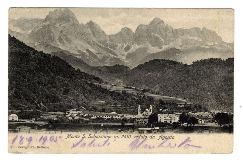 The image depicts a black and white vintage photograph of the Monte S. Sebastiano mountain, located at an elevation of approximately 2410 meters above sea level in Agordo, Italy. The landscape features prominent snow-capped peaks that dominate the background while lush greenery covers lower elevations leading to a village nestled amongst rolling hills.

In the foreground, there is a serene lake reflecting part of the surrounding scenery. A series of buildings and structures can be seen scattered throughout what appears to be a small town or settlement at the base of the mountain range. Among these are several white-domed churches with visible spires, indicating places of worship within this village.

The photograph carries handwritten notations on its lower left side in purple ink that read "1893-1905 Monte S. Sebastiano m. 2410 veduto da Agordo," which translates to "S. Sebastian Mountain at an altitude of 2,410 meters viewed from Agordo." The name P. Breveglieri - Belluno is inscribed in the lower left corner.

Additionally, there are signatures or names written on the right side and bottom right portion of the image that appear to be autographs related to this photograph's provenance or significance at a later time.

The overall atmosphere conveyed by the photo suggests an early 20th-century setting with historical architectural elements reflecting European design.