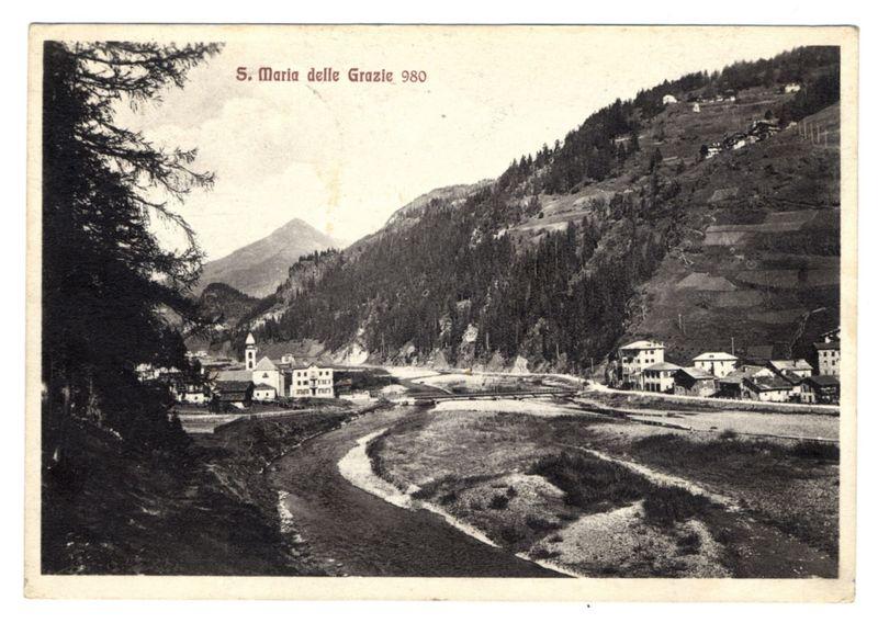 The image is a vintage black and white photograph depicting the village of S. Maria delle Grazie in 1980, as indicated by the text at the top of the photo. The scene shows a picturesque view with mountainous terrain in the background, lush greenery on slopes, and dense forests covering parts of the hillsides.

The foreground features a meandering river or stream flowing through what appears to be farmland or open fields, bordered by patches of bare earth and sparse vegetation. Small settlements are scattered throughout the landscape, consisting mostly of two-story buildings with gabled roofs typical of alpine architecture.

Several structures stand out due to their white facades against the otherwise darker surroundings - a church-like building featuring what seems like a clock tower in the center, as well as other residential or commercial buildings along the river. The village appears sparsely populated and nestled within the natural landscape.