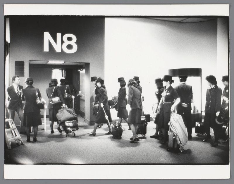 The image captures a busy scene at an airport terminal, with multiple people of various ages and genders standing in line. They appear to be travelers waiting for their flight or luggage collection, as evidenced by the presence of suitcases on wheels. The individuals are dressed primarily in dark colors, suggesting either a uniform dress code typical for airline staff or perhaps just a general trend among passengers at that time.

The airport terminal is labeled with "N8" prominently displayed above the queue line, which might indicate gate N8 within this part of the terminal building. There's also signage indicating it could be an area specifically reserved for stewardesses ("Stewardessen"), as mentioned in the caption from Loener collection and further elaborated on by Stoverman.

The setting is monochromatic with a stark contrast between light and shadow, typical of black-and-white photography which was more prevalent before color photography became widespread. The lighting seems natural or fluorescent, coming perhaps through overhead lights common in airports to illuminate large open spaces efficiently. Notable features include the variety in people's attire and posture as they navigate their way within this transit space.

Overall, it depicts a slice of life at an airport terminal from around 1980-1989, capturing both individual travelers' experiences and possibly pr [...]