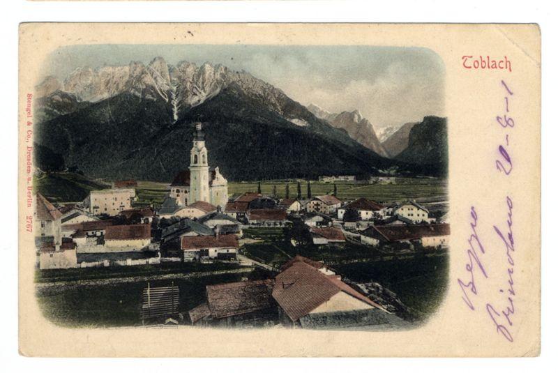 The image shows a vintage postcard depicting an aerial view of the town of Toblach, with prominent alpine mountains in the background. In the foreground is a small village featuring buildings with red-tiled roofs and various architectural styles ranging from simple farmhouses to more ornate structures that could be religious or civic edifices. A notable white building with a spire stands out as it appears larger than other nearby constructions, suggesting its importance within the community.

The mountains behind present an imposing backdrop of rugged peaks, indicating this region is likely situated in a mountainous area such as the Dolomites or another similar range in Europe. The sky above shows subtle gradients from light to darker shades, which could be interpreted either during dawn or dusk lighting conditions.

Overlaying on the upper right corner is red text that reads "Toblach," indicating the name of the town depicted. On the lower left side, there's handwritten inscriptio