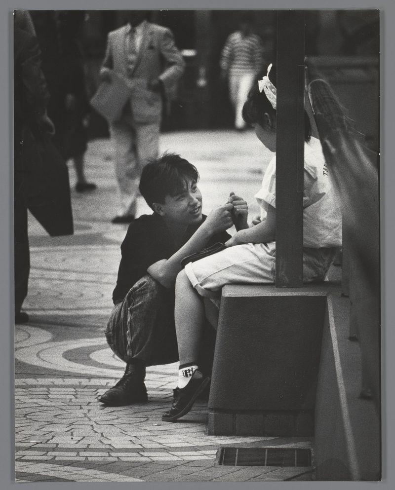 This black and white photograph captures a candid moment on an urban street. In the foreground, two individuals are engaged in what appears to be a close interaction or conversation. The person seated is dressed in dark clothing with distinctive socks bearing text graphics, while their companion stands closer, possibly gesturing towards them.

The background features several pedestrians walking by, adding depth and context to the scene. Their attire suggests a mix of casual and business styles typical for city life. Architectural elements such as paving stones underfoot indicate an urban setting, likely designed with pedestrian traffic in mind.

This image is associated with Ed van der Elsken's work captured during his tenure as Stel op Straat (Stranded on Street) photojournalist in Japan, 1988. The photograph exemplifies a moment frozen in time that reflects the human condition amidst urban life and social interactions.