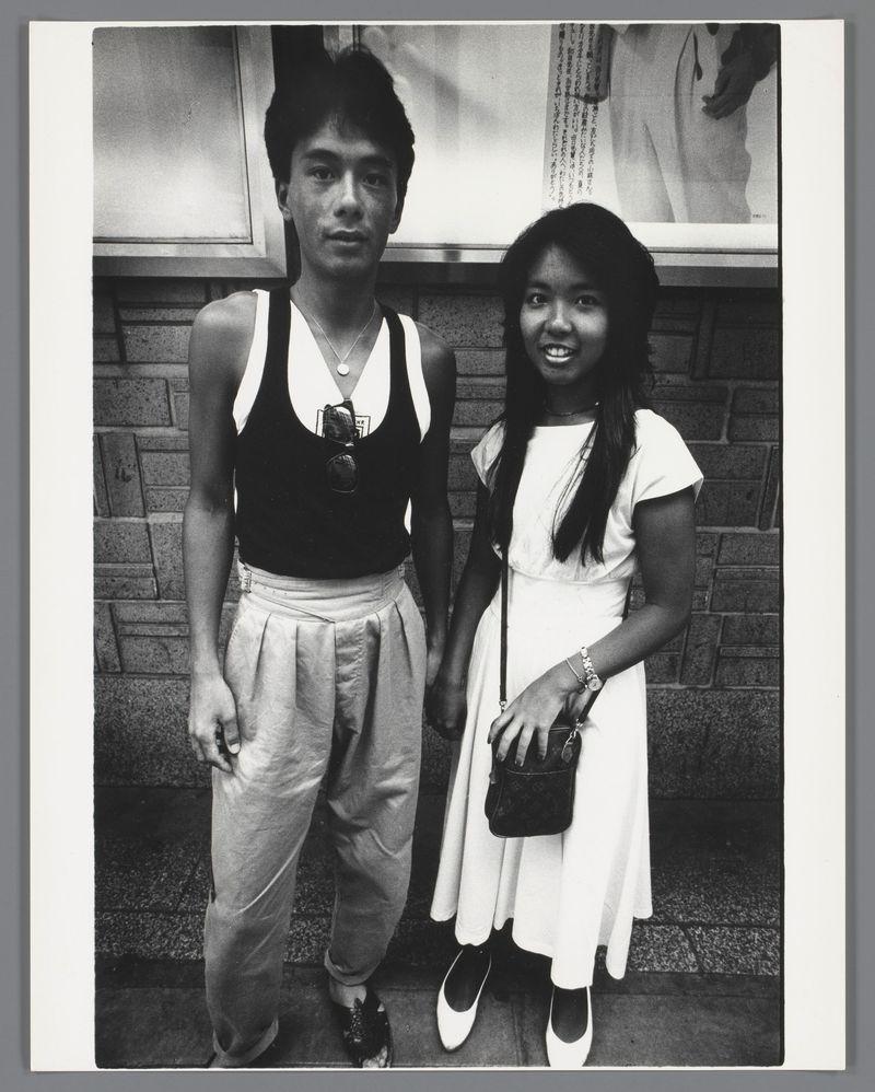 A black and white photograph featuring two individuals standing side by side against a brick wall. On the left, an adult male with dark hair is wearing glasses around his neck on a chain, a sleeveless shirt over a long-sleeve undershirt, high-waisted pants secured at the waist with buttons or clasps, and shoes adorned with ornamental spikes. Beside him stands a young female with shoulder-length black hair, dressed in a white dress with short sleeves, holding a small purse on her right side and wearing flat ballet flats. Both individuals are smiling, suggesting they may be posing for this photograph during an event. Japanese characters can be seen at the top of the image's background, indicating that it might have been taken in Japan or related to Japanese culture or subjects. The caption 'Portret van een onbekend jong stel' suggests a casual portrait with unidentified youth.