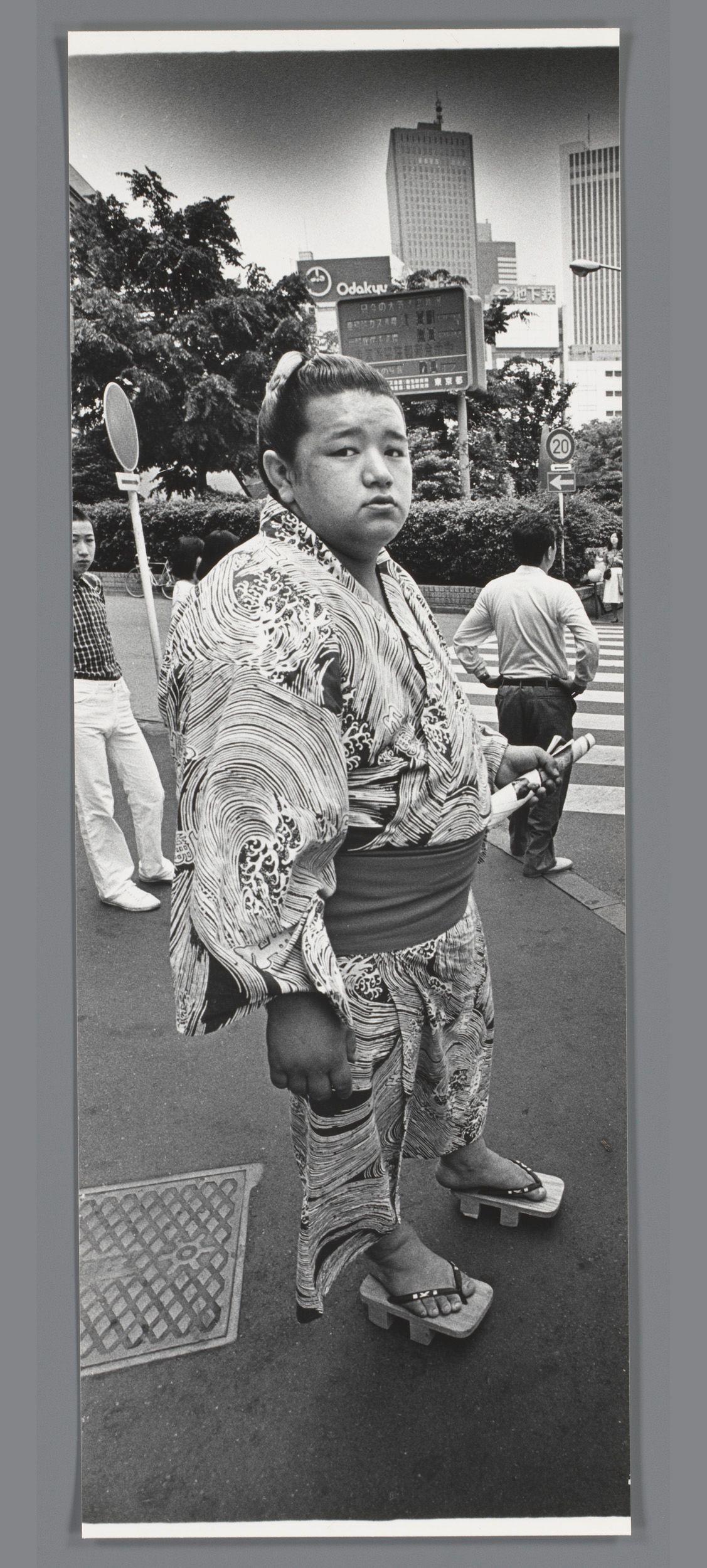 The image is a black and white photograph capturing an individual dressed in traditional Japanese sumo wrestling attire, known as "maikobo," on a city street. The person stands prominently at the center of the frame, facing slightly to their right with a neutral expression. They are wearing a patterned kimono that has swirling designs predominantly in shades of black and white.

The surroundings suggest an urban setting during daylight hours; there's another individual walking away from the camera on the right side, while others can be seen further back in the scene. The backdrop includes trees, signage for "Odakyu," indicating a public transportation hub or nearby station, traffic lights, pedestrian crossing signs with numbers, and various buildings that give an impression of a modern city environment.

Notable features include traditional Japanese sandals on wooden platforms known as geta, which are visible at the bottom. The overall composition captures a moment juxtaposing cult