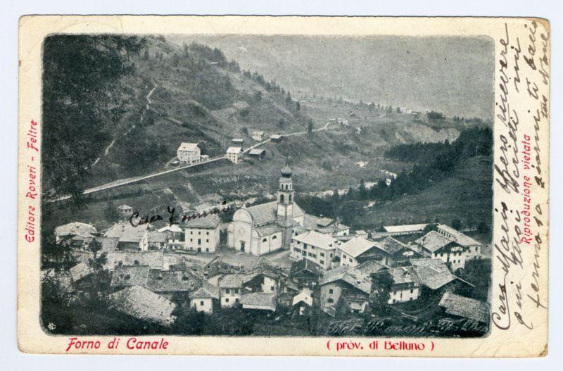 This image is a vintage postcard depicting the village of Forno di Canale, which was part of Belluno province. The photograph captures an aerial view of the settlement nestled within hilly terrain. Prominent in the center is a church with twin bell towers, indicative of its importance to community life. Surrounding buildings vary in architecture and size but are primarily clustered around what appears to be main thoroughfares that traverse through town. Some structures have tiled roofs while others seem more austere or industrial. The landscape suggests rural characteristics, possibly indicating the time period's economic reliance on agriculture or traditional crafts like iron working suggested by "Forno," which translates as furnace in Italian, hinting at possible historical industry significance within this locality. No people are visible to infer demographic details such age groups or gender ratios. Notable features include handwritten annotations and a printed label that adds conte