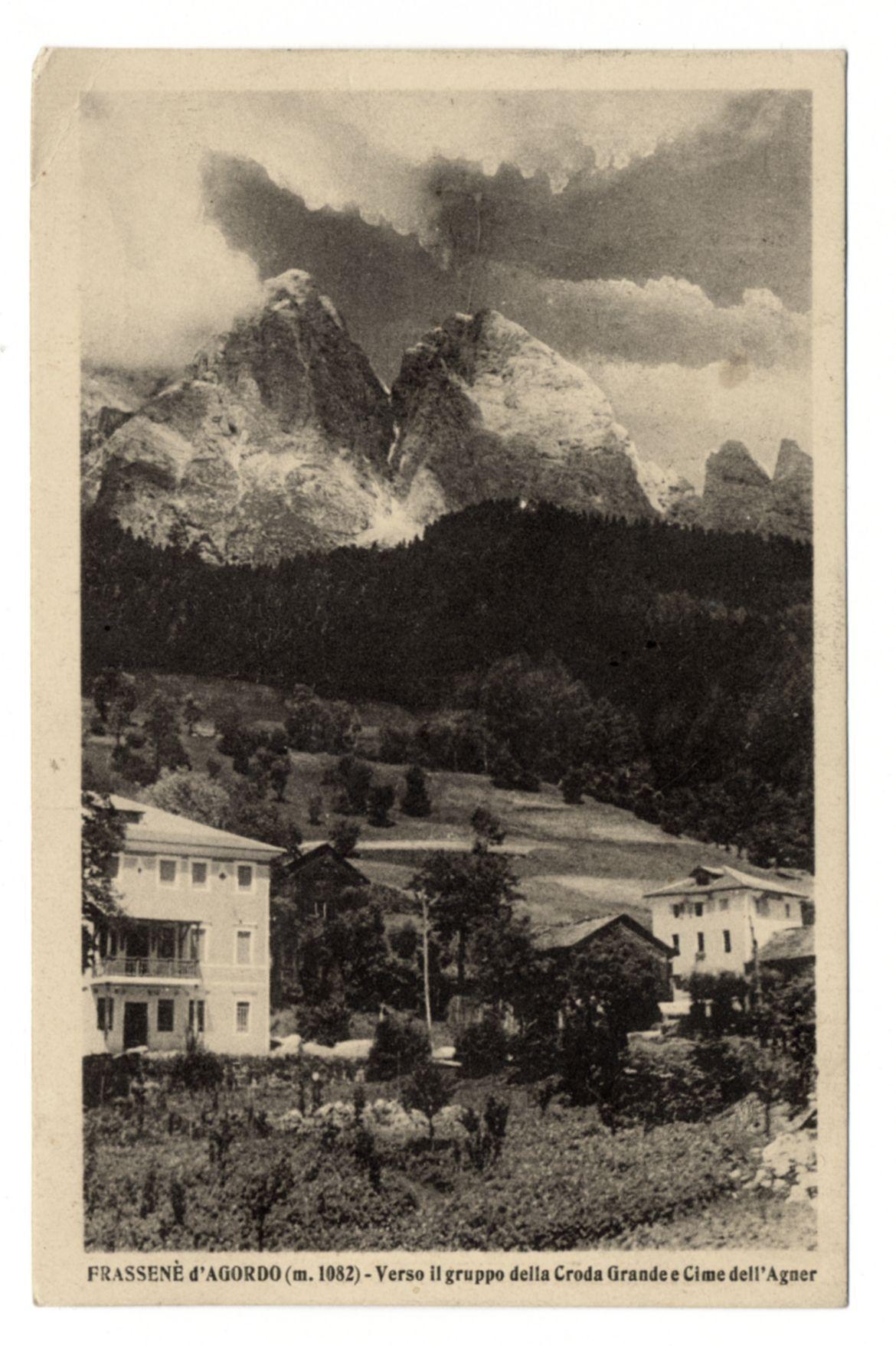 The image depicts a vintage black and white photograph of a mountainous landscape, likely taken in the early 20th century given its style. It features imposing snow-capped peaks rising above dense forests and residential buildings at their base. The foreground shows two-story houses with flat roofs, one prominently to the left and another partially visible on the right side. In front of these structures is a cultivated garden area filled with bushes or small plants.

The central part of the image reveals rugged mountain terrain blanketed in snow, indicating cold weather conditions typically found at high altitudes. The peaks have sharp ridges, some parts are steep while others display gentle slopes leading down to forested regions. A cloudy sky looms overhead, adding a dramatic effect and possibly suggesting variable weather.

Text on the bottom of the image reads "FRASSENE d' AGORDO (m. 1082) - Verso il gruppo della Croda Grande e Cime dell'Agnèr," which translates to "Frasene di 