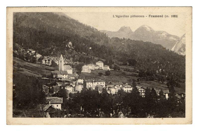 The image is a black and white photograph depicting an old village nestled in the hills, possibly taken from a higher vantage point. It appears to be an aged postcard or print with text at the top indicating "L'Agordino pittorese - Frassanè (m. 1082)" which suggests it is located near L'Agnone Agordino and has an elevation of approximately 3,576 feet. The landscape features dense clusters of buildings typical of a European alpine town with pitched roofs and white facades. A prominent church tower stands out amidst the houses, hinting at its significance in community life.

The background reveals mountainous terrain with hints of snow or mist, suggesting this village is situated within a high-altitude region, possibly in Italy's Dolomite Alps given the style of architecture and landscape resemblance to other Alpine villages such as those found on Ferro Candilera. The text below indicates it was taken around 1908.

The image has signs of wear with creases and discoloration typical of