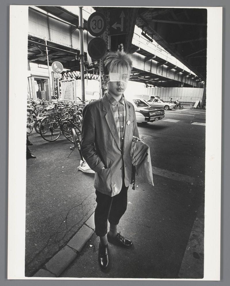 The image is a black and white photograph of an individual standing under an elevated train track. The person appears to be wearing a blazer, checkered shirt, trousers, socks pulled up just above the ankle, and polished shoes. Their hair stands out in spikes or disarray atop their head. They are holding what seems like documents or papers in one hand. In the background, we can observe multiple bicycles parked on both sides of the street, indicating a possible urban setting with cycling infrastructure. A traffic sign is visible above them displaying "30" and an arrow pointing upwards to the right, alongside another circular symbol that suggests pedestrian access through bicycle lanes. Other vehicles are partially seen in the distant background under the bridge structure. The overall environment appears busy yet organized typical of city life.