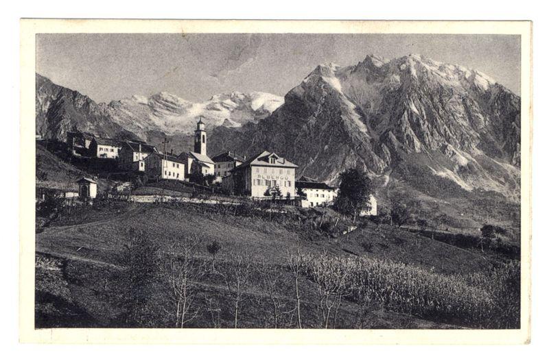 The image is a black and white photograph depicting a small village nestled in the foothills of mountainous terrain. In the foreground, there are barren trees indicating it may be late autumn or early spring. The middle ground shows cultivated fields with varying shades of brown soil and patches that could suggest different types of crops or fallow land.

The background is dominated by tall mountains covered in snow at higher elevations, suggesting a cool climate typical of high-altitude regions. In the center stands a prominent building labeled "ALBERGH," likely indicating it as an inn or hotel for travelers passing through this area.

Surrounding buildings appear to be residential homes and smaller structures that could serve various community purposes such as shops or workshops. The architecture is simple with pitched roofs, which are practical in snowy regions due to their ability to shed snowfall effectively.

The sky above the mountains appears clear without visible clouds, implying fair weather at the time of capture. Overall, this image conveys a peaceful and isolated rural setting, possibly in an alpine region known for its natural beauty and tourism.