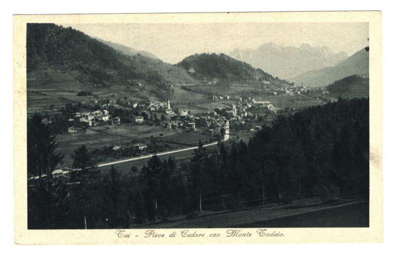 The image is a monochromatic vintage postcard depicting a picturesque landscape. It shows an expansive view of Pieve di Cadore with Monte Tudaio in the backdrop, situated within a valley surrounded by rolling hills and densely wooded areas. The foreground features a paved road winding through greenery leading to clusters of buildings that make up the town or village. Notable structures include what appears to be historical or religious edifices standing out amidst more modest dwellings.

The terrain is varied with patches of cultivated fields, indicating agricultural activity in this rural setting. In the distance, faint outlines suggest a mountain range under an overcast sky. The postcard bears text at its bottom which likely denotes the location and perhaps serves as a title or caption for the photograph.

There are visible signs of wear on the edges of the card, indicative of age and handling through time. This vintage appeal adds to the historical context of the image, hinting that it captures a scene from past times in this region. The overall composition provides an insight into rural life during the period when such postcards were popular as keepsakes or for sharing far-flung places with distant loved ones.