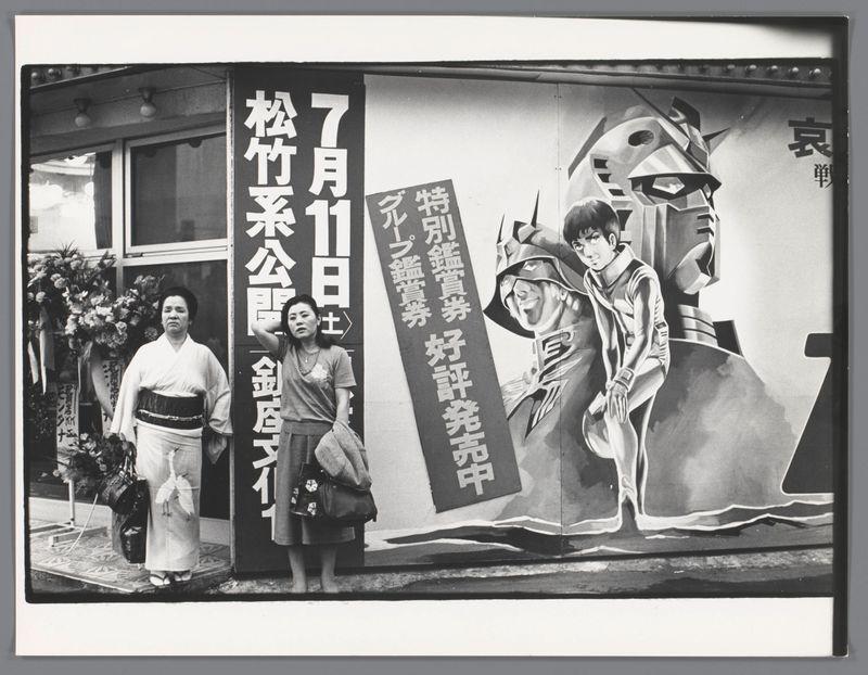 The image shows a street scene in Japan with two women standing next to an advertisement painted on the side of a building. The woman on the left is dressed in traditional Japanese attire, including a kimono and geta sandals, while the woman on the right wears modern clothing and carries bags. Behind them is a large mural featuring illustrations from what appears to be a comic or graphic novel with text written in Japanese characters.

The advertisement includes prominent white lettering against a black background that reads "July 10" followed by more Japanese script, suggesting an event or announcement related to the date mentioned. The artwork on the mural depicts three figures: one character is wearing futuristic armor and helmet, while another appears as a soldier with a hat; these characters are likely from a sci-fi theme given their stylized design.

The scene captures everyday life juxtaposed against popular culture elements, providing insight into contemporary Japanese society during that period. The photograph was taken by Ed van der Elsken in 1982 and is part of his body of work exploring various aspects of urban Japan's cultural landscape.