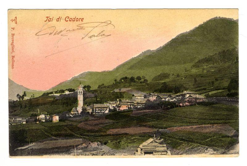The image displays a vintage postcard with the title "Tai di Cadore" prominently displayed at the top in cursive script, suggesting an Italian origin. The word appears twice: once larger and centered near the top of the card, followed by a signature or inscription beneath it which is less legible but seems to be another personal mark possibly indicating authorship or dedication.

The background depicts a scenic landscape dominated by rolling hills with varying shades of green indicative of vegetation or crops, likely vineyards. The terrain appears hilly and terraced in certain areas, typical of agricultural land use for planting vines or other plants that require specific elevation levels.

In the foreground, there are clusters of buildings clustered together; these structures vary slightly but generally resemble traditional European rural architecture with red roofs and pale walls. A prominent feature is a bell tower rising above the cluster on the left side, suggesting this may b