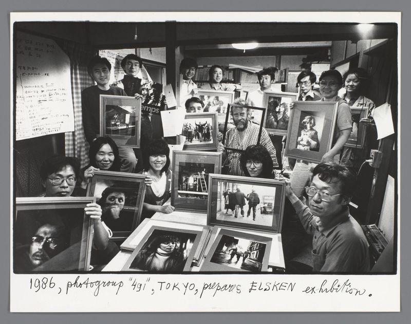 The image is a black and white photograph featuring a group of people, possibly photographers or artists, gathered together in what appears to be an indoor setting with shelves and items that resemble books or files. They are holding framed photographs, suggesting they may have been involved in some form of photography exhibition or project.

In the center foreground, there's a man wearing glasses looking directly at the camera, his expression somewhat neutral but attentive. He is surrounded by others who seem to be smiling or focused on their frames. The people vary widely in age and style; both men and women are present, with different hairstyles indicating diverse tastes or perhaps eras.

The setting looks cluttered yet organized, indicative of a workspace for creative individuals where ideas might flow freely among the group members. There's an air of casual professionalism about them as they showcase their work to others. The lighting is even across most faces, though some are