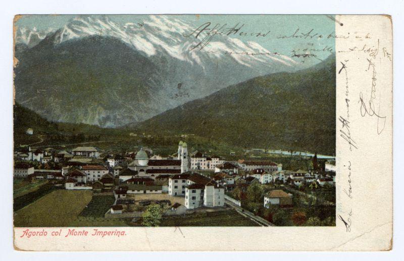 The image depicts a picturesque, vintage postcard showing an aerial view of Agordo with Monte Imperina in the background. This mountainous landscape is characterized by snow-capped peaks and lush green valleys, creating a stark contrast between earthy tones below and crisp whites above.

In the foreground lies the town itself, displaying an array of buildings showcasing historical architecture typical of European alpine regions. Prominent structures include what appears to be a large monastery or abbey with distinctive domes and spires standing out amidst other residential edifices. The color palette is rich but muted, suggesting age; sepia tones dominate, evoking nostalgia.

Handwritten annotations in the upper right corner add a personal touch, potentially indicating ownership or sentiment associated with this particular postcard. Below it, additional writing confirms the location as "Agorado col Monte Imperina," reinforcing its identity and setting for viewers familiar with Ital