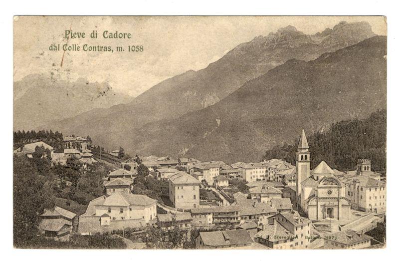 The image is a sepia-toned photograph of an old town nestled in the mountains, labeled "Pieve di Cadore dal Colle Contras m. 1058." The text suggests that this could be Pieve di Cadore taken from Colle Contras at approximately 3429 feet above sea level. In the foreground are various buildings with pitched roofs and European architectural styles indicative of a small, historical town. Prominent among these is an older church or cathedral with distinctive features such as its bell tower rising higher than surrounding structures. The background reveals steep mountain slopes covered in dense vegetation, leading up to more rugged peaks. This landscape conveys the setting's remote nature within a valley surrounded by mountains.