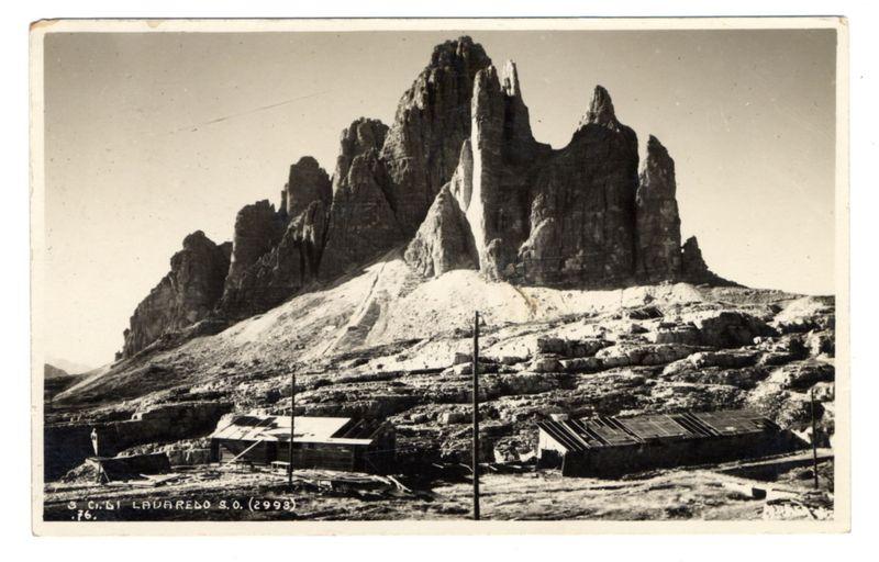 The image is a black and white photograph depicting a barren landscape with tall, jagged rock formations rising dramatically against the sky. These rocks appear weathered and have pointed peaks that suggest erosion over time. In the foreground of the composition, there are several dilapidated structures: two small buildings or sheds to the left, one with an open roof exposing its interior contents, and a larger structure on the right side which appears more stable but is also showing signs of age such as peeling paint and what might be rusted metal parts. Between these shelters stands a solitary tall pole, possibly a telephone or telegraph post. The ground around the buildings is uneven with some debris scattered about, indicating that this area may not have been maintained for years. There's minimal vegetation visible, which reinforces the arid conditions of the environment. A handwritten inscription at the bottom reads "3.61 Lavaredo S.O. (2998)" followed by a signature and possibly 