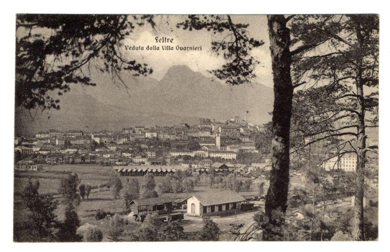 The image is a black and white vintage photograph, likely from the early 20th century. It depicts an expansive view of Feltre as seen from Villa Guarnieri. In the foreground are trees with dense foliage on either side framing the picture. The middle ground shows a rural landscape leading up to more urbanized areas, including several buildings and what appears to be train tracks or railway lines curving into the distance.

The background reveals a densely populated town with multiple structures ranging from smaller houses to larger institutional-looking edifices. A prominent tower rises above many of these buildings, suggesting it could be an important landmark in Feltre's skyline. Beyond the urban sprawl is a mountain range that looms over the landscape, adding depth and grandeur.

The text "Seltre Veduta dalla Villa Guarnieri" at the top suggests this photograph captures Seltre as seen from Villa Guarnieri. The overall composition offers an atmospheric snapshot of Feltre's geograp