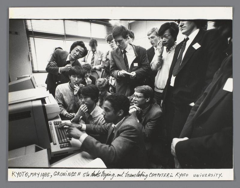 The image is a black and white photograph capturing a group of people gathered around an older model computer, which appears to be from the early 1980s or late 1970s. The setting seems to be a classroom or lab within Kyoto University in Japan during May 1985. Multiple individuals are standing and sitting closely together, with some looking intently at the screen of the computer as they engage in what appears to be collaborative work or study.

The group is diverse, including both men and women wearing suits, business casual attire, and more casual clothing like a grey sweatshirt. Many have name tags on their lapels indicating that this might be an organized event such as a conference or workshop involving students from Groningen University in the Netherlands visiting Kyoto University for academic purposes.

The atmosphere is one of focused attention and collaboration among participants. The presence of various age groups suggests different levels of experience with technology, whic