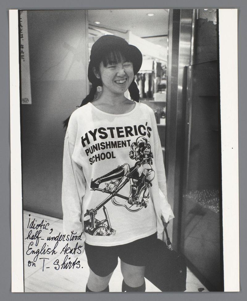 The image shows a young girl standing in an indoor setting, possibly on the second floor of a building given her proximity to what appears to be part of an elevator's exterior. The girl is smiling and closing one eye as if she’s squinting playfully or winking at the camera. She wears a wide-brimmed hat with bangs that cover her forehead completely, dark hair peeking out from beneath it in braids pulled back into what looks like ponytails on either side of her head.

She is dressed casually; wearing short black shorts and knee-high socks paired with sneakers. A white long-sleeved T-shirt stands out due to its distinctive print - a stylized illustration that reads "HYSTERICS' PUNISHMENT SCHOOL" above an image depicting what appears to be a person on roller skates or similar inline skating equipment, holding onto something resembling handles of the rollerskating apparatus.

The photograph is in black and white, which gives it a timeless quality. The top edge has handwritten notes that