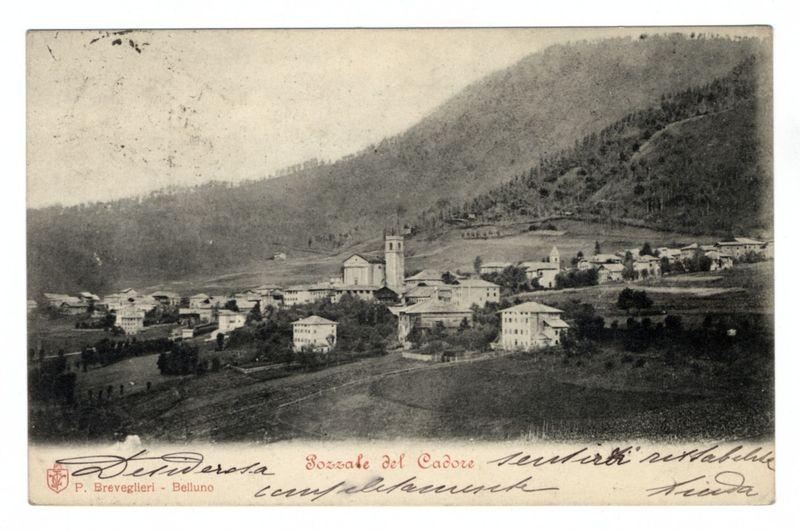 The image is a black and white photograph depicting an old, small town nestled in the foothills of a hill or mountain. It appears to be historical due to its aged appearance with faded edges at some points and handwritten script on it which adds authenticity.

Several buildings are visible; these include what seems like residential homes scattered across the hillsides as well as larger structures that could serve communal purposes, such as schools or town halls. Prominently in view is a bell tower rising above several other buildings near the center of the image, indicating its possible importance within this community - perhaps serving both religious and possibly administrative functions.

The landscape surrounding the settlement shows gentle slopes with sparse trees and vegetation, typical for hilly regions. The sky is not visible; however, it seems to be overcast as indicated by the lack of strong shadows or bright highlights in the image.

At the bottom left corner, there's a